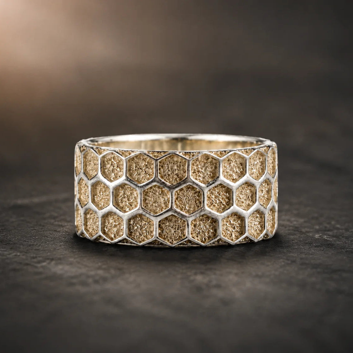 A wide silver ring featuring a textured honeycomb pattern filled with gold-toned sand, displayed on a dark textured surface.