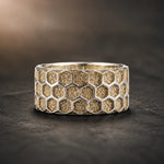 A wide silver ring featuring a textured honeycomb pattern filled with gold-toned sand, displayed on a dark textured surface.