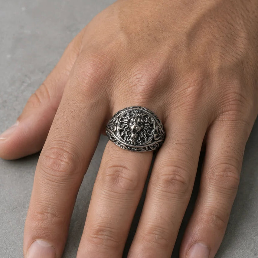 A silver-toned filigree signet ring with a detailed floral or scrollwork design, shown worn on a person's ring finger against a textured gray background.