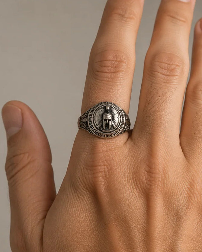 A silver signet ring with an engraved circular emblem worn on the ring finger of a hand, shown against a neutral gray background.