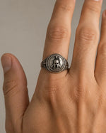 A silver signet ring with an engraved circular emblem worn on the ring finger of a hand, shown against a neutral gray background.