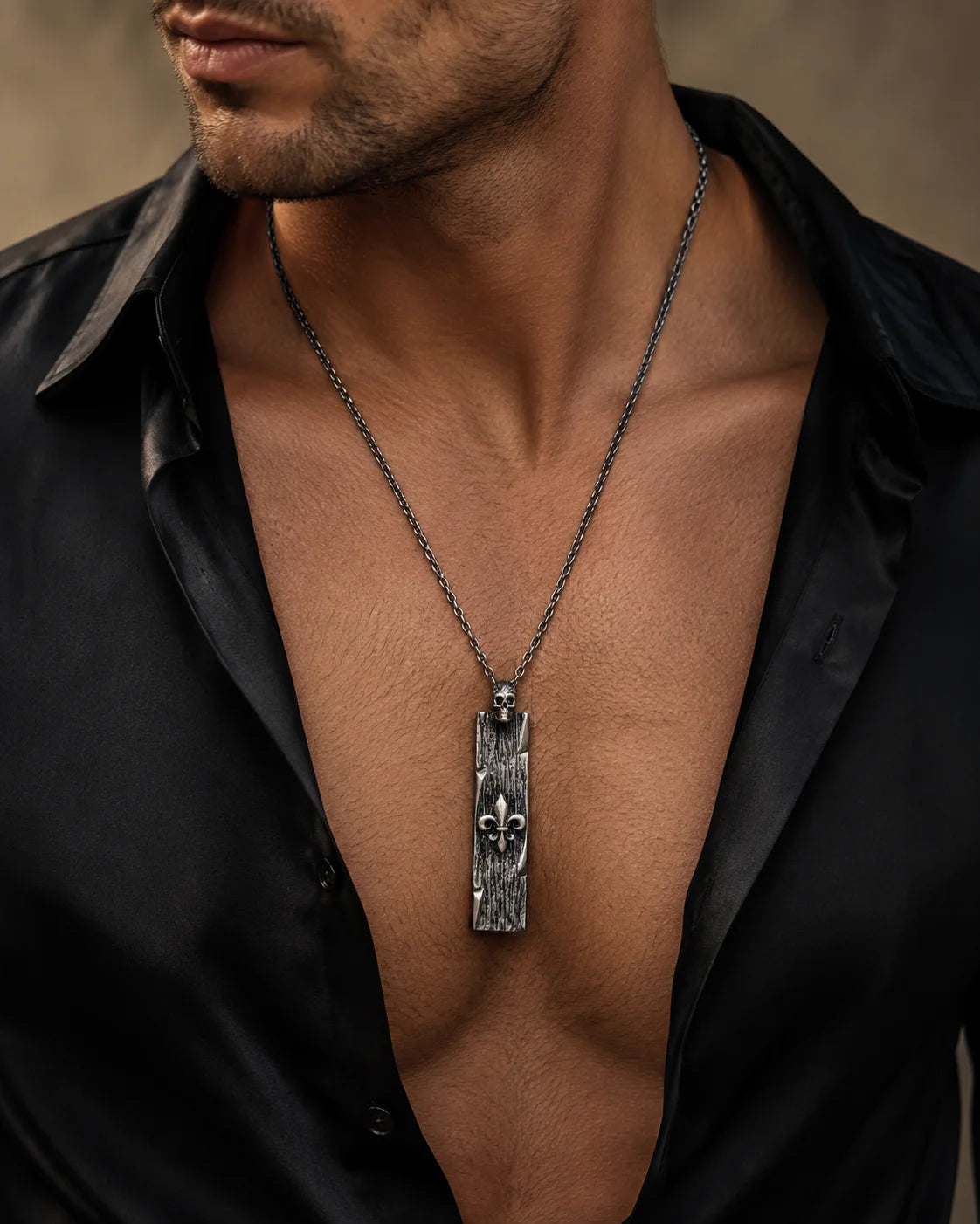 A close-up of a man wearing a dark metal pendant necklace with a textured rectangular bar and fleur-de-lis design, paired with an open black shirt.