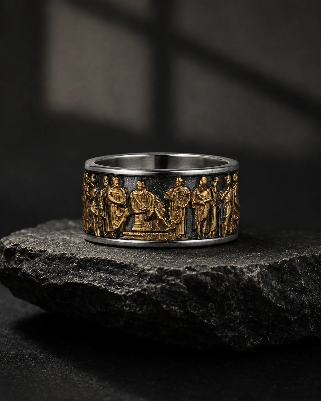 A wide silver-toned ring with raised gold-toned figures and a central seated figure, displayed on a dark textured stone against a dark moody background.
